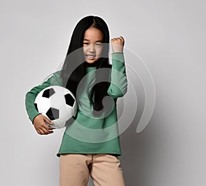 Happy Asian kid girl holding soccer football ball in one hand and gesturing Yes, Done with the other hand