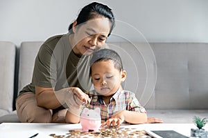 Happy Asian kid and mom saving money together, putting cash into piggy bank. Mother playing with child on heating floor at home,