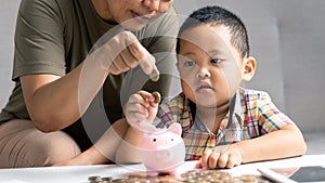 Happy Asian kid and mom saving money together, putting cash into piggy bank. Mother playing with child on heating floor at home,