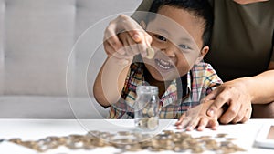 Happy Asian kid and mom saving money together, putting cash into piggy bank. Mother playing with child on heating floor at home,