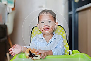 Happy Asian little baby boy sitting on children chair indoor eating bread with Stuffed Chocolate-filled dessert and Stained around