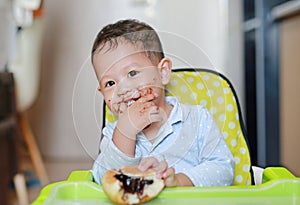 Happy Asian little baby boy sitting on children chair indoor eating bread with Stuffed Chocolate-filled dessert and Stained around