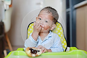 Happy Asian little baby boy sitting on children chair indoor eating bread with Stuffed Chocolate-filled dessert and Stained around