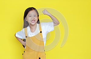 Happy asian little child girl showing his muscle with looking camera isolated on yellow background with copy space. Strong kid in