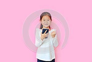 Happy asian little child girl watching her smartphone and expression hand fight sign over pink background. Kid smiling and enjoy