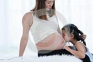 Happy Asian little cute kid girl playing with pregnant mom on bed at home, touch and kiss mother belly with tender and love.