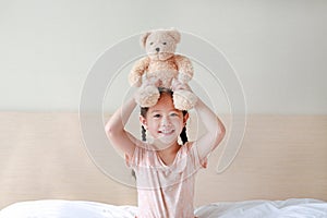 Happy Asian little girl Picking up brown teddy bears on her head while sitting on the bed at home