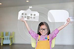 Happy Asian little girl in pink-yellow dungarees holding mock-up paper school and empty blank speech bubble to tell something in