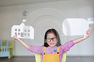 Happy Asian little girl in pink-yellow dungarees holding mock-up paper school and empty blank speech bubble to tell something in