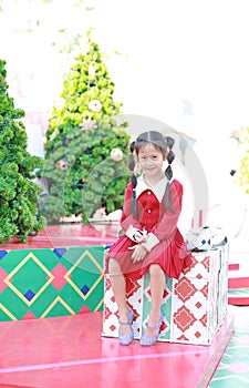 Happy Asian little girl in red dress sitting on big gift box against Christmas tree in a background
