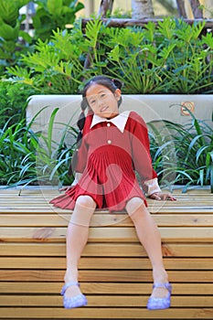 Happy Asian little girl in red dress sitting and lying on wood bench in the garden