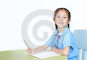 Happy Asian little girl in school uniform writing on notebook at desk isolated on white background. Schoolgirl and Education