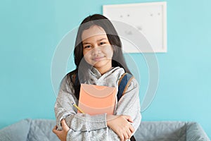 Happy Asian little girl smile on the face, Asia child hold book and backpack while standing in interior home. Portrait of a kid