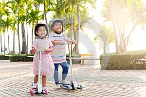 Happy Asian little kid boy and girl wear safe helmet play kick board on road in park outdoors on summer day