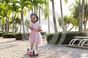 Happy Asian little kid girl wear safe helmet playing pink kick board on road in park outdoors on