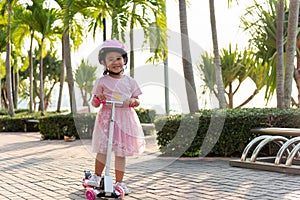 Happy Asian little kid girl wear safe helmet playing pink kick board on road in park outdoors on summer day