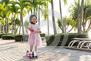 Happy Asian little kid girl wear safe helmet playing pink kick board on road in park outdoors on summer day