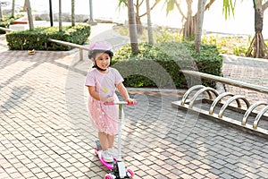 Happy Asian little kid girl wear safe helmet playing pink kick board on road in park outdoors on summer day