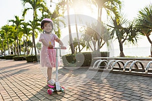 Happy Asian little kid girl wear safe helmet playing pink kick board on road in park outdoors on summer day