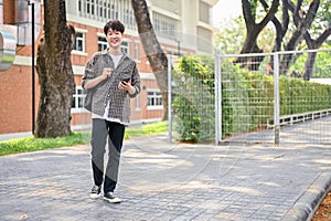 A happy Asian male college student is using his smartphone while walking in the campus park