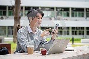 A happy Asian male college university student listening to music in a campus park alone