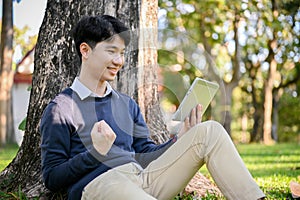 Happy Asian male using his tablet in the park, showing clenched fist, receiving good news