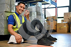 Happy Asian male warehouse worker in uniform sitting in warehouse, using walkie-talkie