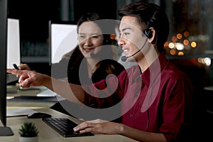 Happy Asian man with headphones work night shift at call center customer care service desk with colleagues, point at computer