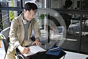 Happy Asian man making lists on his personal planner book, remote working at the coffee shop