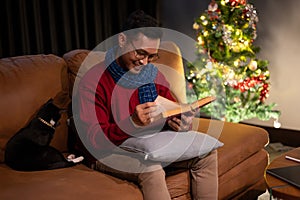 A happy Asian man is reading a book on a sofa while his cat is sitting beside him, Christmas at home