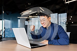 A happy Asian man sits in the office with laptop, talks on a video call, rejoices at the success