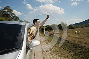 Happy Asian man in sunglasses leaning out from car window against blue sky. Travel and transportation concept