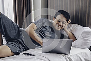 Happy Asian man is working with his laptop on his bed. Concept of freelancer successful lifestyle