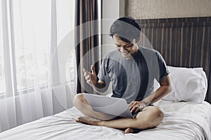 Happy Asian man is working with his laptop on his bed. Concept of freelancer successful lifestyle