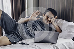 Happy Asian man is working with his laptop with video call in a bed. Concept of freelancer successful lifestyle