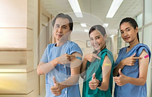 Happy Asian medical personnel in uniform stand pointing and show bandage at shoulder after vaccine injection in hospital