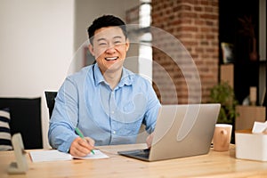 Happy asian middle aged man working on laptop and taking notes in notebook, smiling at camera, free space