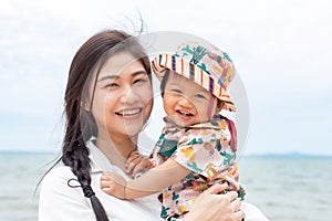 Happy Asian mom and baby smile looking at camera with beautiful sea. Vacation of family mother and baby spending time together