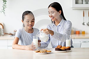 Happy asian mom and daughter eating snacks and drinking milk in kitchen interior, having a bite together