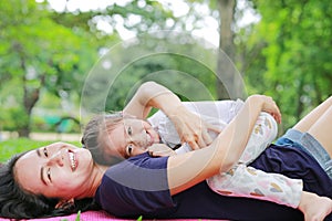Happy Asian mom embrace her daughter lying in the green garden with looking camera. Funny mother and child girl playing in summer