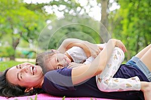 Happy Asian mom embrace her daughter lying in the green garden with looking camera. Funny mother and child girl playing in summer