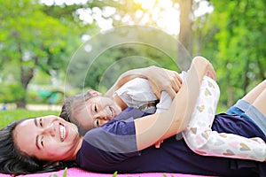 Happy Asian mom embrace her daughter lying in the green garden with looking camera. Funny mother and child girl playing in summer