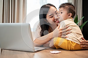 A happy Asian mom is playing and feeding her little son with snacks while working in her home office