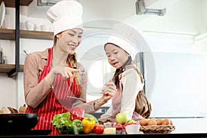 Happy Asian mother and daughter girl wear apron chef hat at kitchen, mom touch transparent device tablet for searching tasty meal