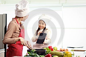 Happy Asian mother daughter wear apron and chef hat, mom hold cooking tasty fried meal with pan at kitchen, look at digital tablet