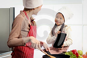 Happy Asian mother daughter wear apron and chef hat, mom hold cooking tasty fried meal with pan at kitchen, look at digital tablet