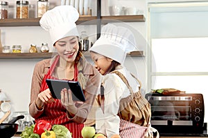 Happy Asian mother daughter wearing apron and chef hat at kitchen, mom and girl kid look at digital tablet for searching tasty