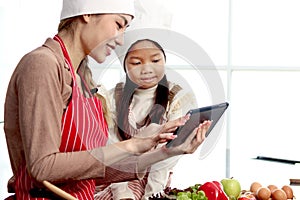 Happy Asian mother daughter wearing apron and chef hat at kitchen, mom and girl kid look at digital tablet for searching tasty