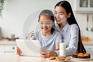 Happy asian mother and her daughter using digital tablet, looking for new recipes while sitting at kitchen, free space