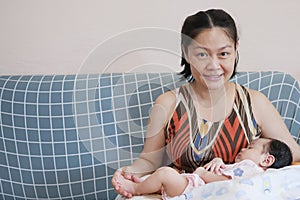 Happy Asian mother holding her sleeping baby, sitting on sofa